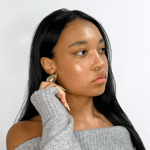 Woman wearing Alvéra Gold Bold Waterdrop Stud Earrings with gray off-shoulder sweater – elegant gold statement studs, classy everyday winter jewelry.